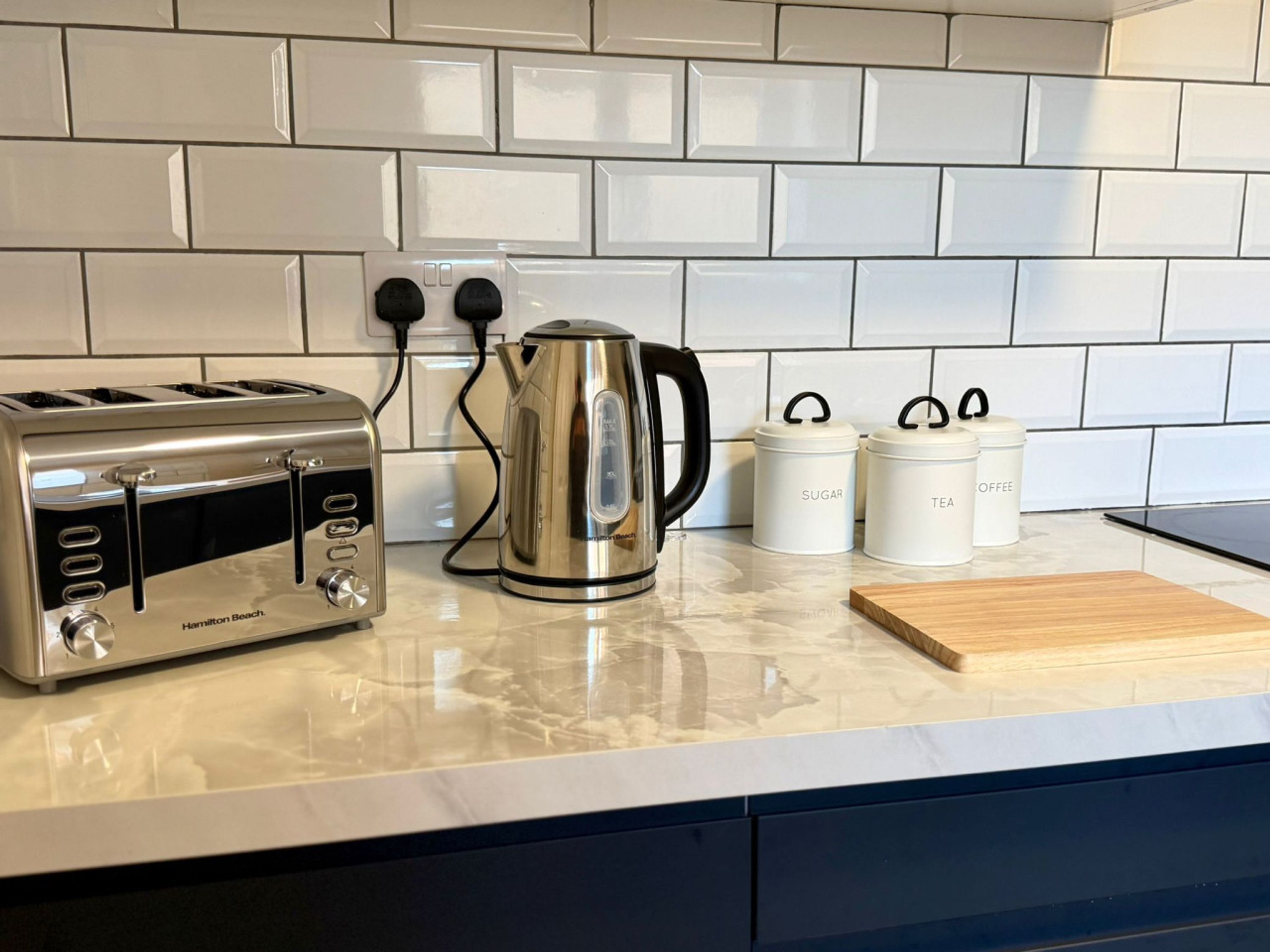 A kitchen with a Hamilton Beach toaster, a stainless steel kettle, and white storage containers labeled 'SUGAR' and 'TEA'.