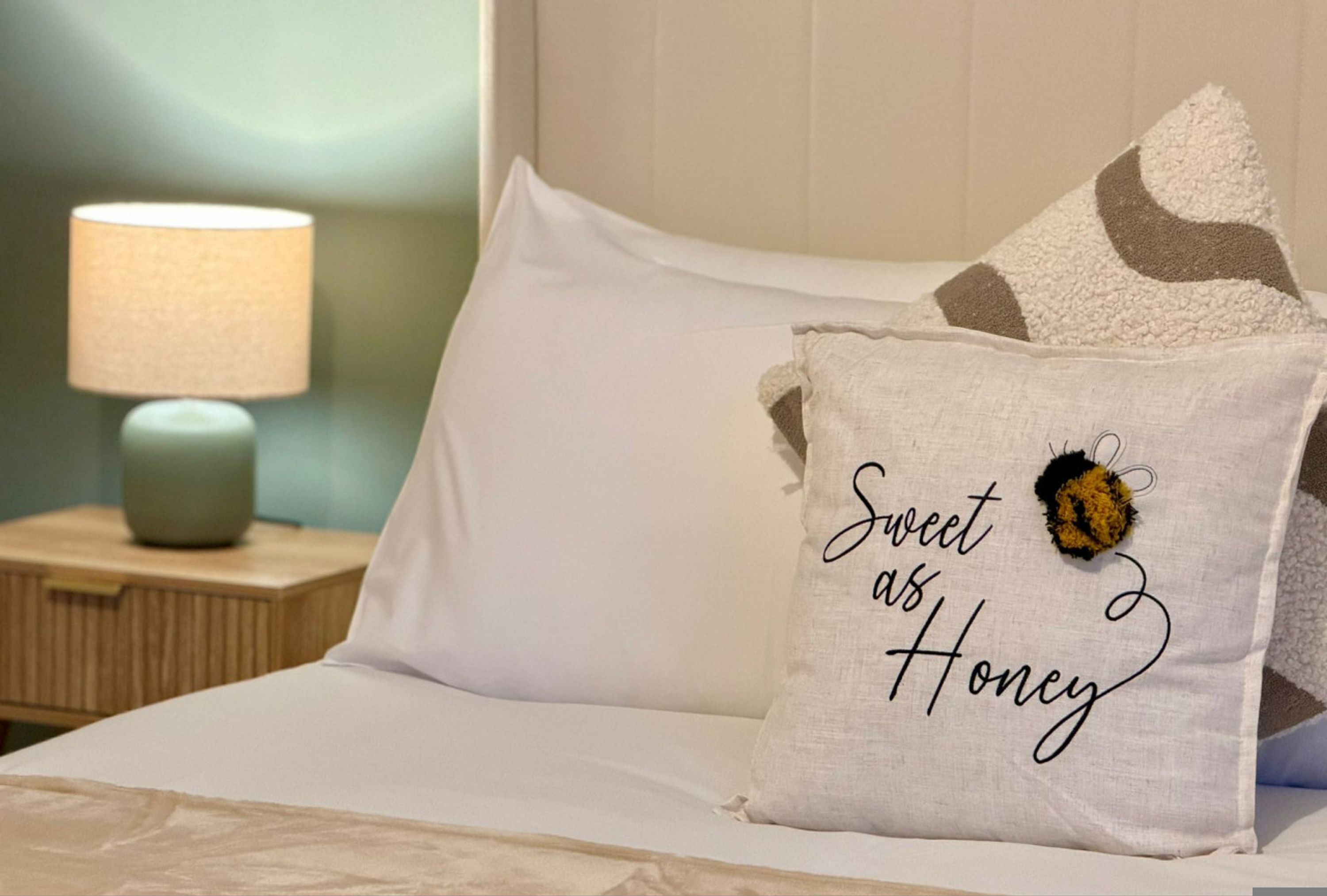 A bed with white bedding, a pillow with 'Sweet as Honey' text, a bee decoration, and a bedside lamp.