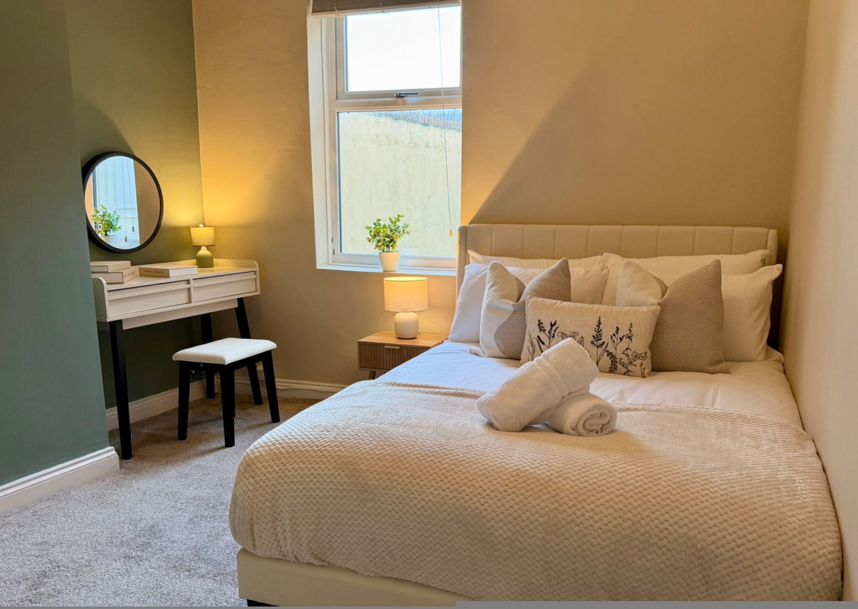 A bedroom with a bed, a bedside table with a lamp, a mirror, a desk, and a chair.