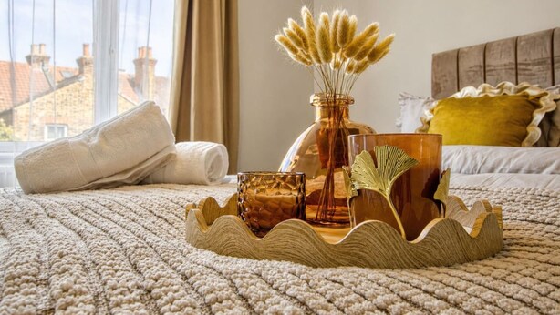 Una cama con una bandeja decorativa, un florero con plumas de pampa y un vaso sobre una colcha con textura.