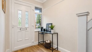 Un pasillo con una puerta blanca, una ventana con vistas, una mesa consola de metal negro con una planta y una cesta.