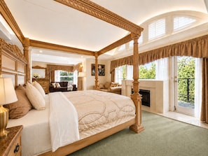 A four-poster bed with a green carpet, a fireplace, and a balcony with a view of greenery.