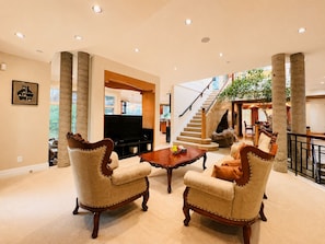 A spacious living room with a large TV, two upholstered chairs, a wooden coffee table, and a staircase leading to an upper level.