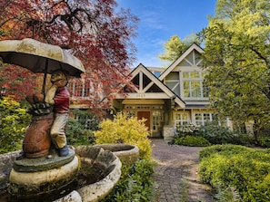 A statuary fountain with a figure holding an umbrella, a house with a number '3489' on the facade, and a brick pathway leading to the entrance.