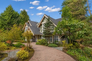 A two-story house with a brick pathway leading to the entrance, surrounded by lush greenery and trees.
