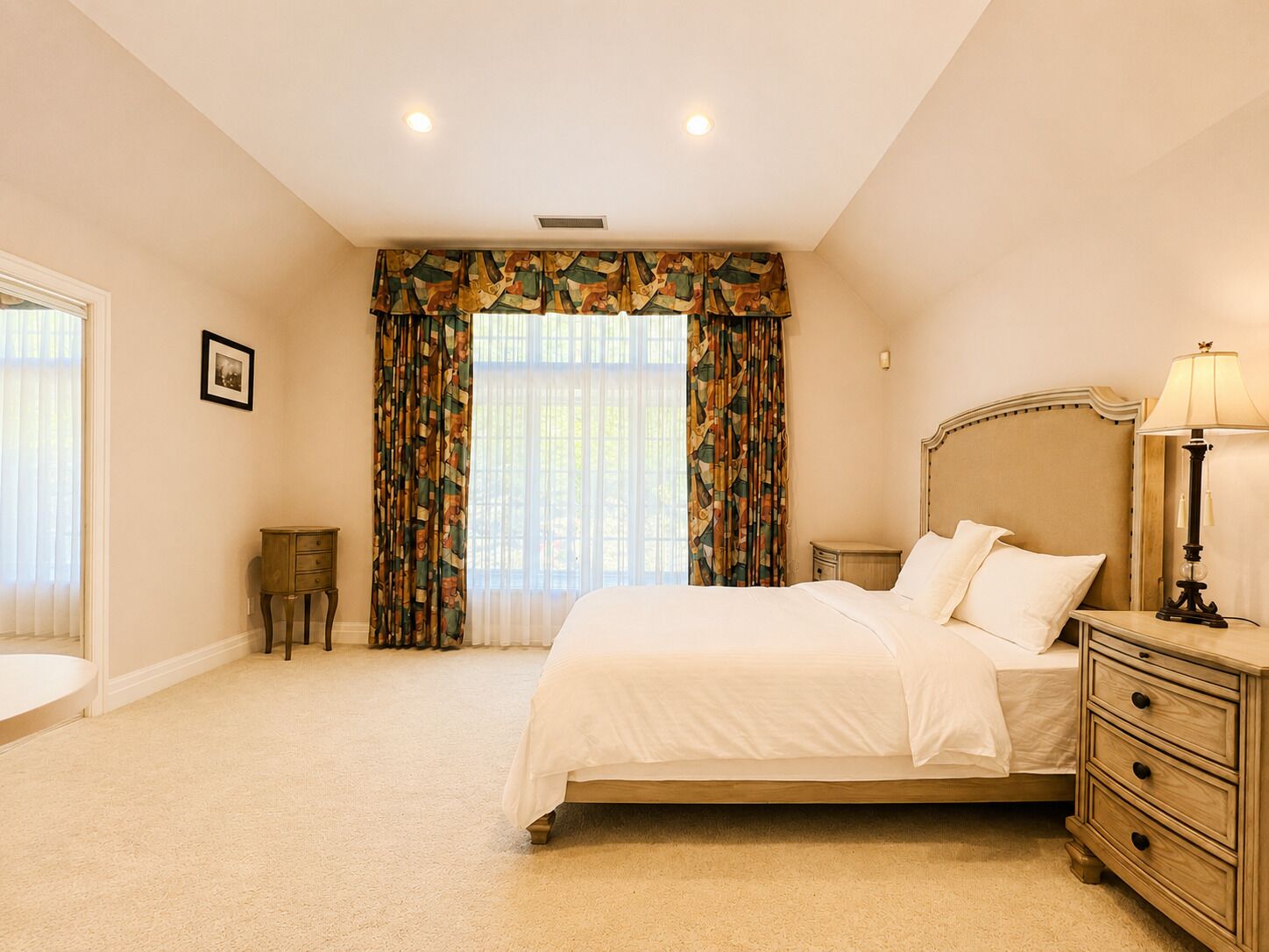 Une chambre à coucher avec un lit, une table de chevet, une lampe, des rideaux et une petite table.