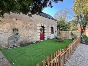 Ein Steinhaus mit roter Tür, ein kleiner Garten mit Holzzaun und ein Tisch mit Stühlen.