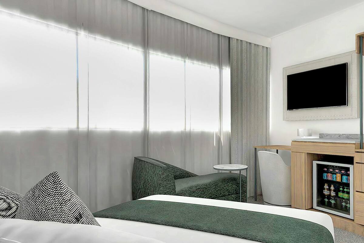Moderni hotellihuone, jossa on suuri sänky, työpöytä, jossa on televisio, minibaari ja suuri ikkuna, jossa on verhot.