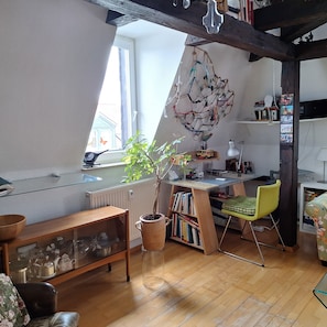 Ein gemütliches Zimmer mit schrägen Wänden, einem Holztisch, einem grünen Stuhl, einem Heizkörper und einem Fenster mit Blick auf ein Haus.