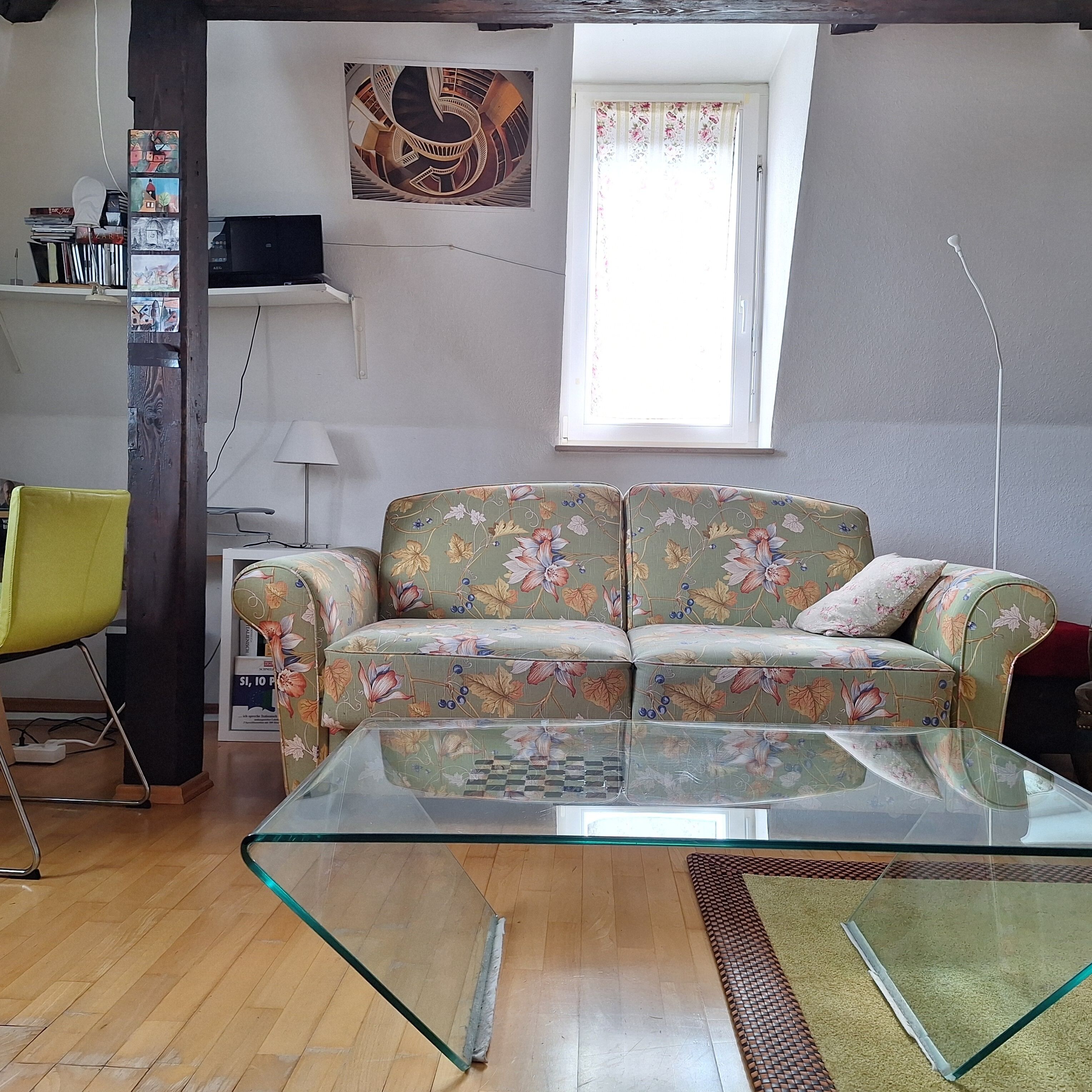 Ein Wohnzimmer mit einem blumigen Sofa, einem Glastisch, Holzboden, einem Fenster mit floralen Vorhängen und einem an der Wand hängenden Kunstwerk.