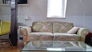 Ein Wohnzimmer mit einem blumigen Sofa, einem Glastisch, Holzboden, einem Fenster mit floralen Vorhängen und einem an der Wand hängenden Kunstwerk.