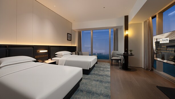 A hotel room with two beds, a desk, a chair, a view of the city, and a large window. - ChangZhou DaJiuDian (Changzhou)