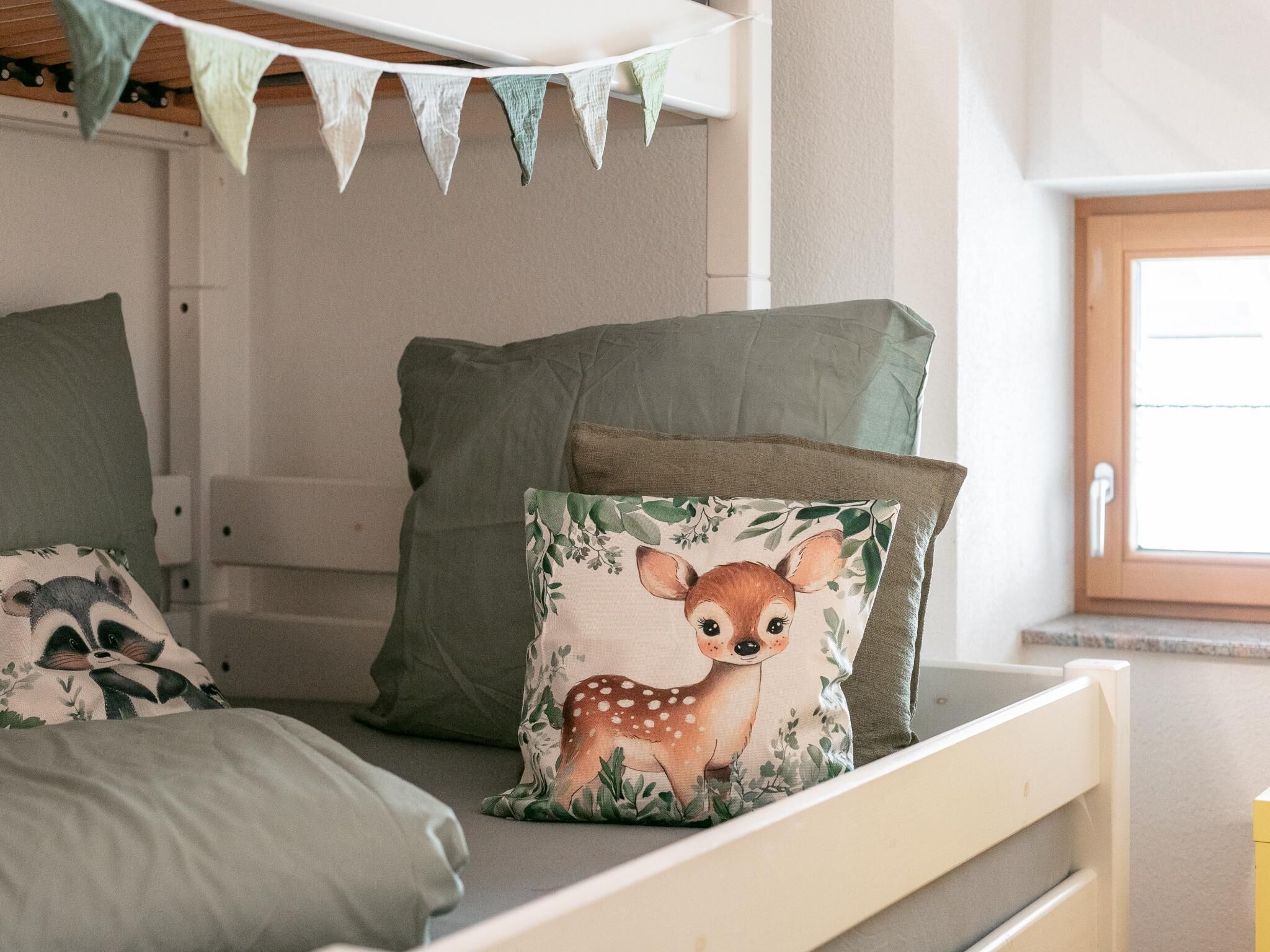 Un lit superposé avec une literie verte, un coussin décoratif orné d’un motif de cerf et une fenêtre offrant une belle vue.