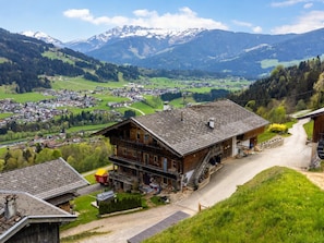 Il s’agit d’un bâtiment traditionnel en bois, doté d’un toit en pente, d’un escalier apparent menant à l’entrée et offrant une vue sur une vallée avec des maisons et des montagnes en arrière-plan.