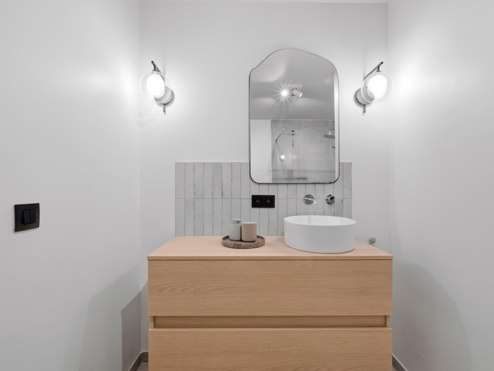Een moderne badkamer met een houten wastafelmeubel, een rond wastafel, een spiegel en twee wandlampen.