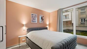 A bedroom with a bed, a small bedside table, a wall-mounted light, and a window with a view of buildings.