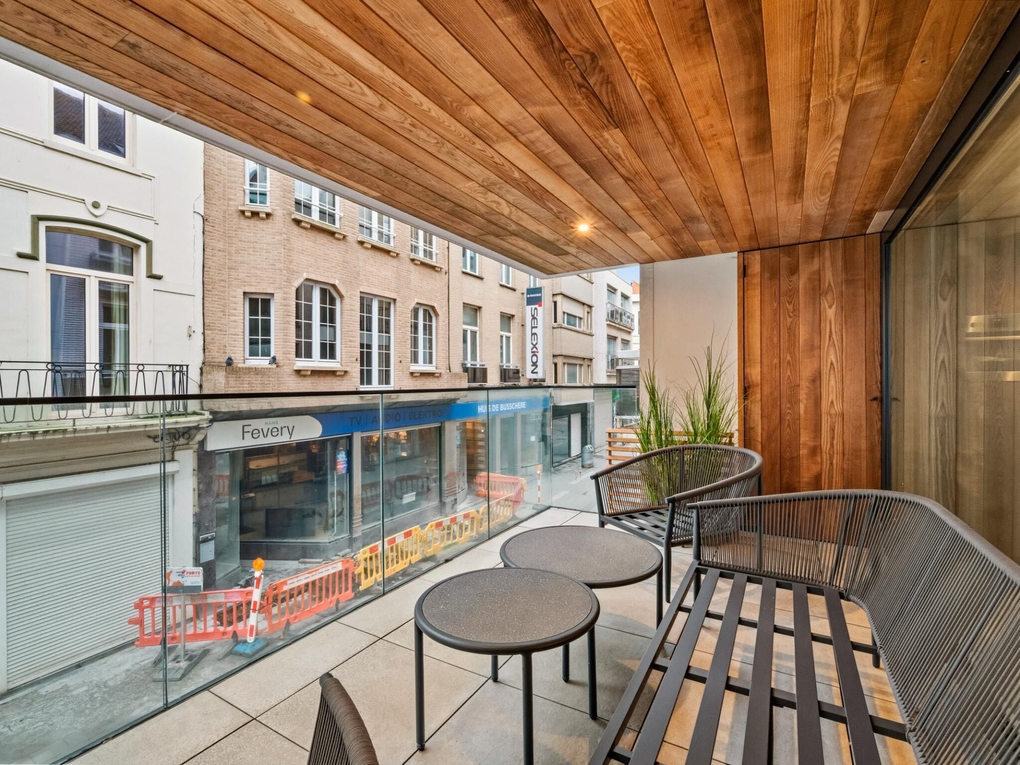 A modern outdoor seating area with a wooden ceiling, a view of the street, and a glass barrier.