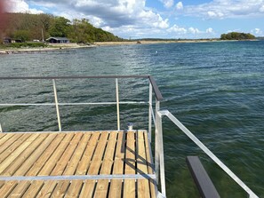 Une jetée en bois s’étendant dans une étendue d’eau, avec une rive lointaine où l’on aperçoit des bâtiments et des arbres.
