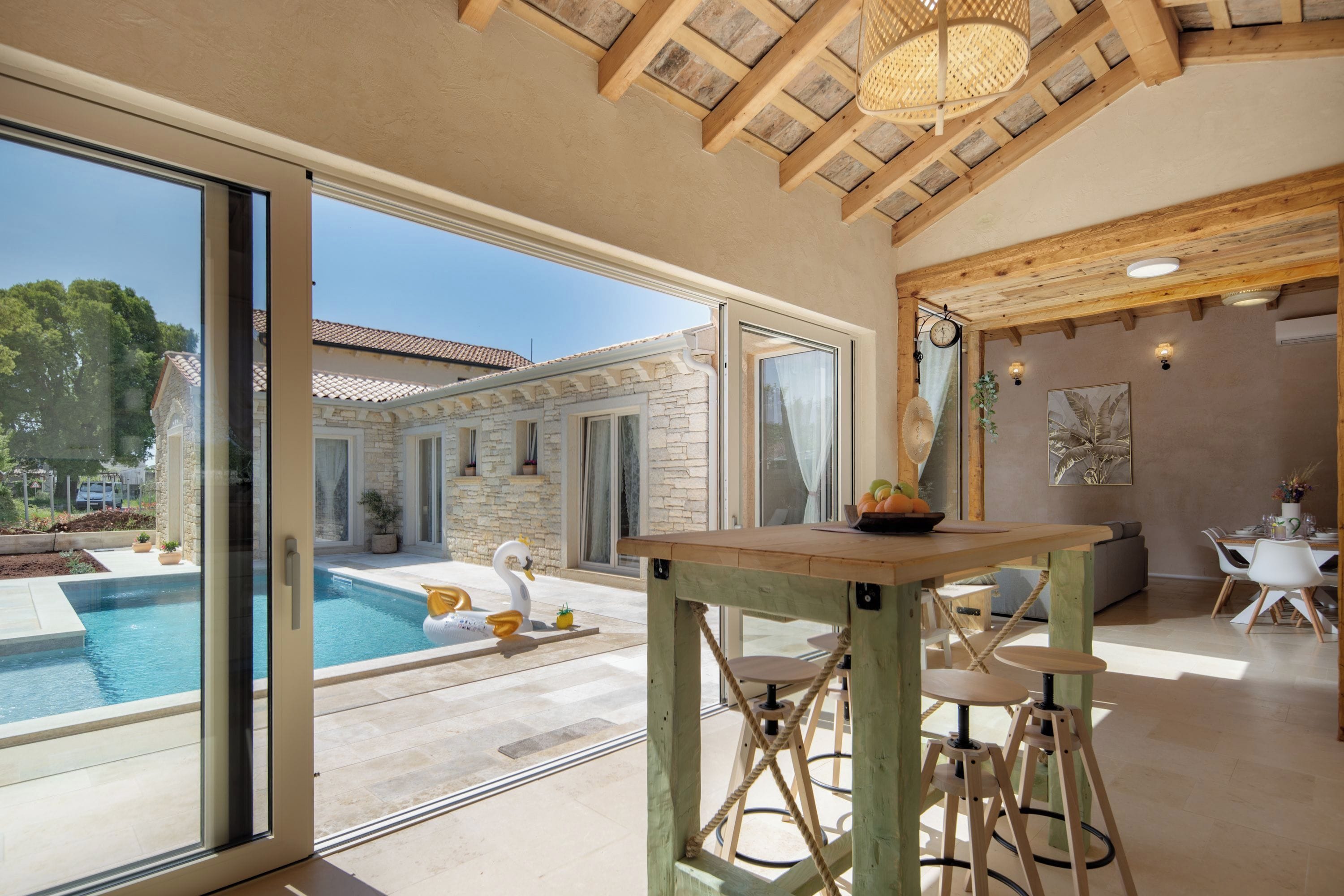 Un interior moderno con vista a la piscina, vigas de madera en el techo y un comedor con taburetes.