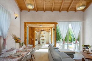 A spacious room with a high ceiling, wooden beams, a dining area with a table set for four, a sofa, and a view of a garden through large windows. - Villa Stonica - Rustic Istrian Pool Retreat (Tinjan)