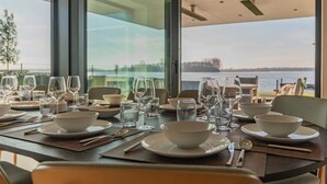 Tavola imbandita con piatti, ciotole, bicchieri e posate, con vista su uno specchio d'acqua attraverso ampie finestre.