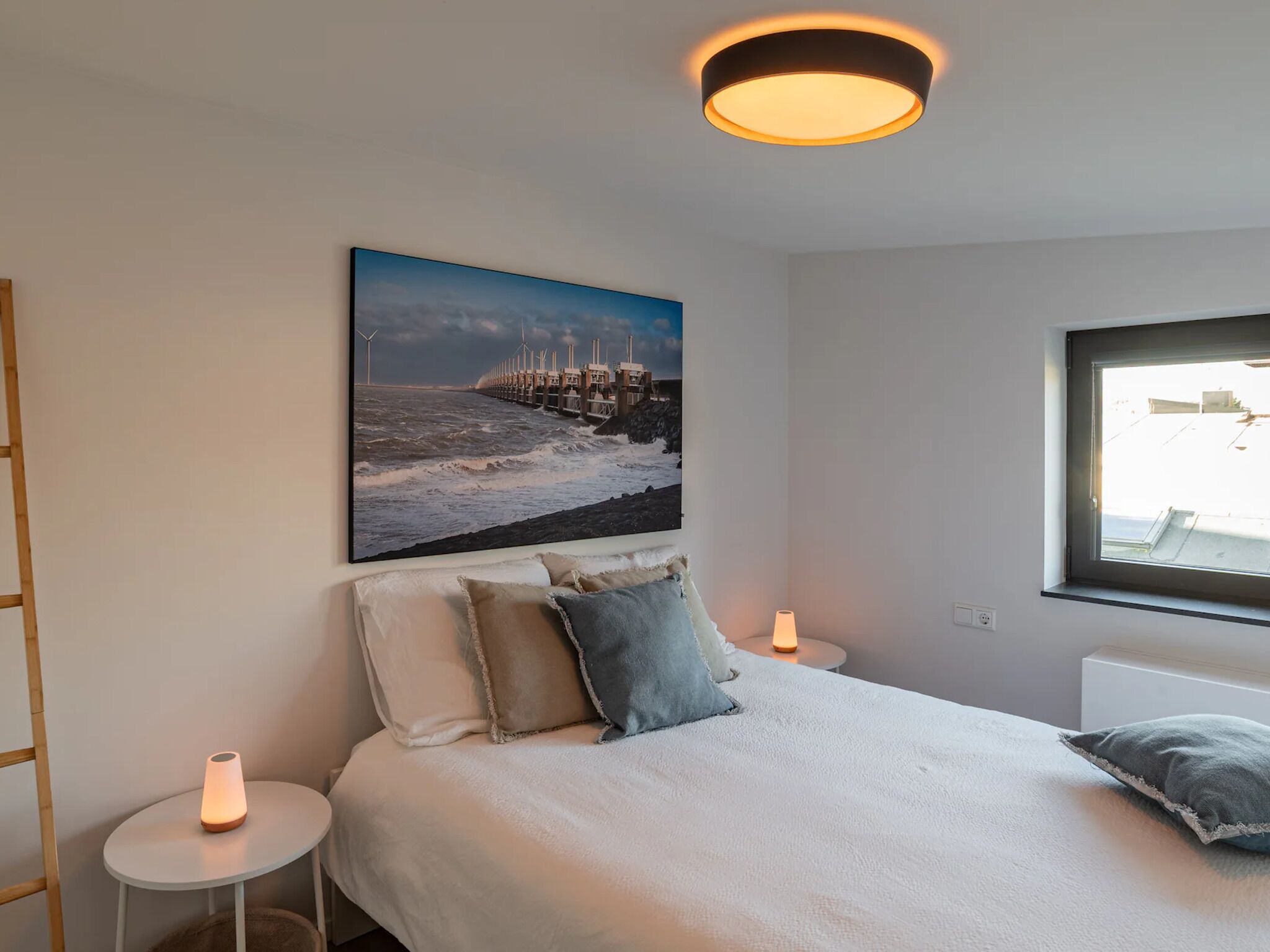 A bedroom with a bed, pillows, a bedside table with a lamp, a window, and a painting of a coastal scene.