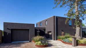 Un edificio moderno a due piani con garage, un giardino anteriore curato con piante e un cielo azzurro intenso.