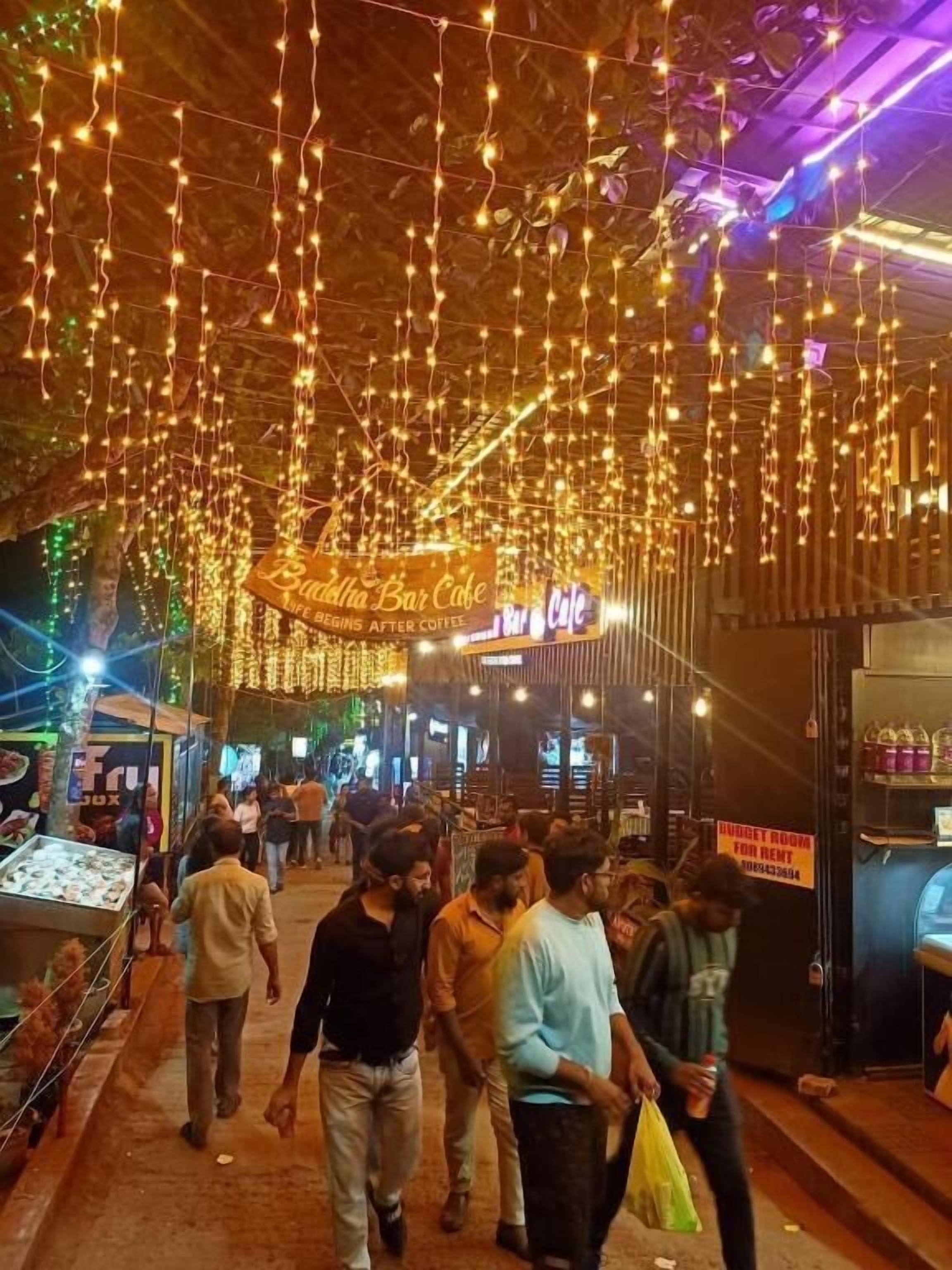 A crowded indoor market with string lights, a sign for 'Buddha Bar Cafe,' and a 'Budget Room for Rent' sign.