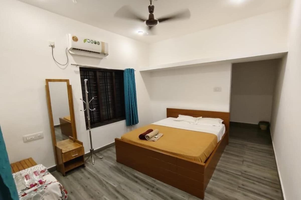A bedroom with a bed, a ceiling fan, an air conditioning unit, a window with curtains, a mirror, and a small table.