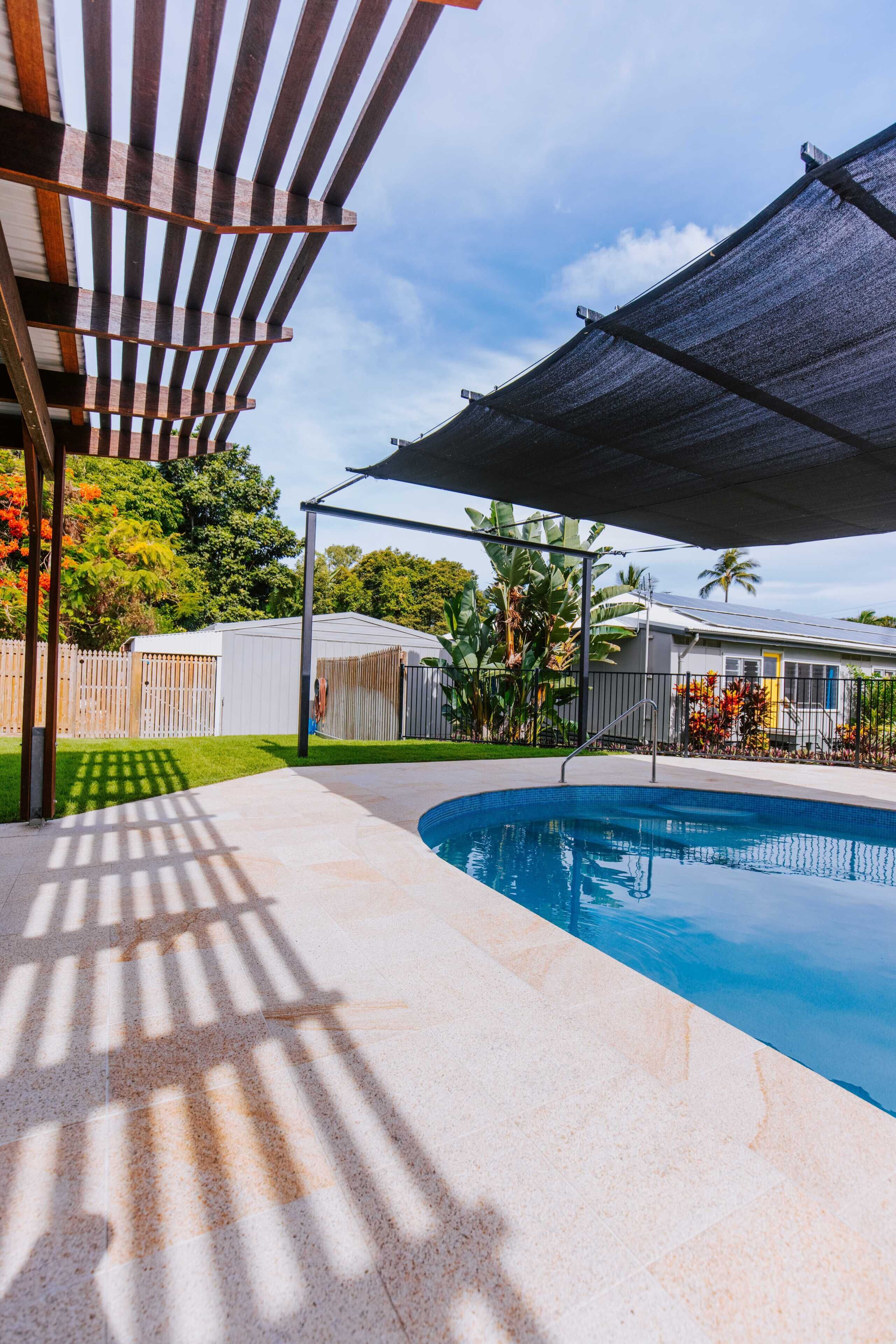 Una piscina con una pérgola y zona sombreada, rodeada por una valla y vegetación.