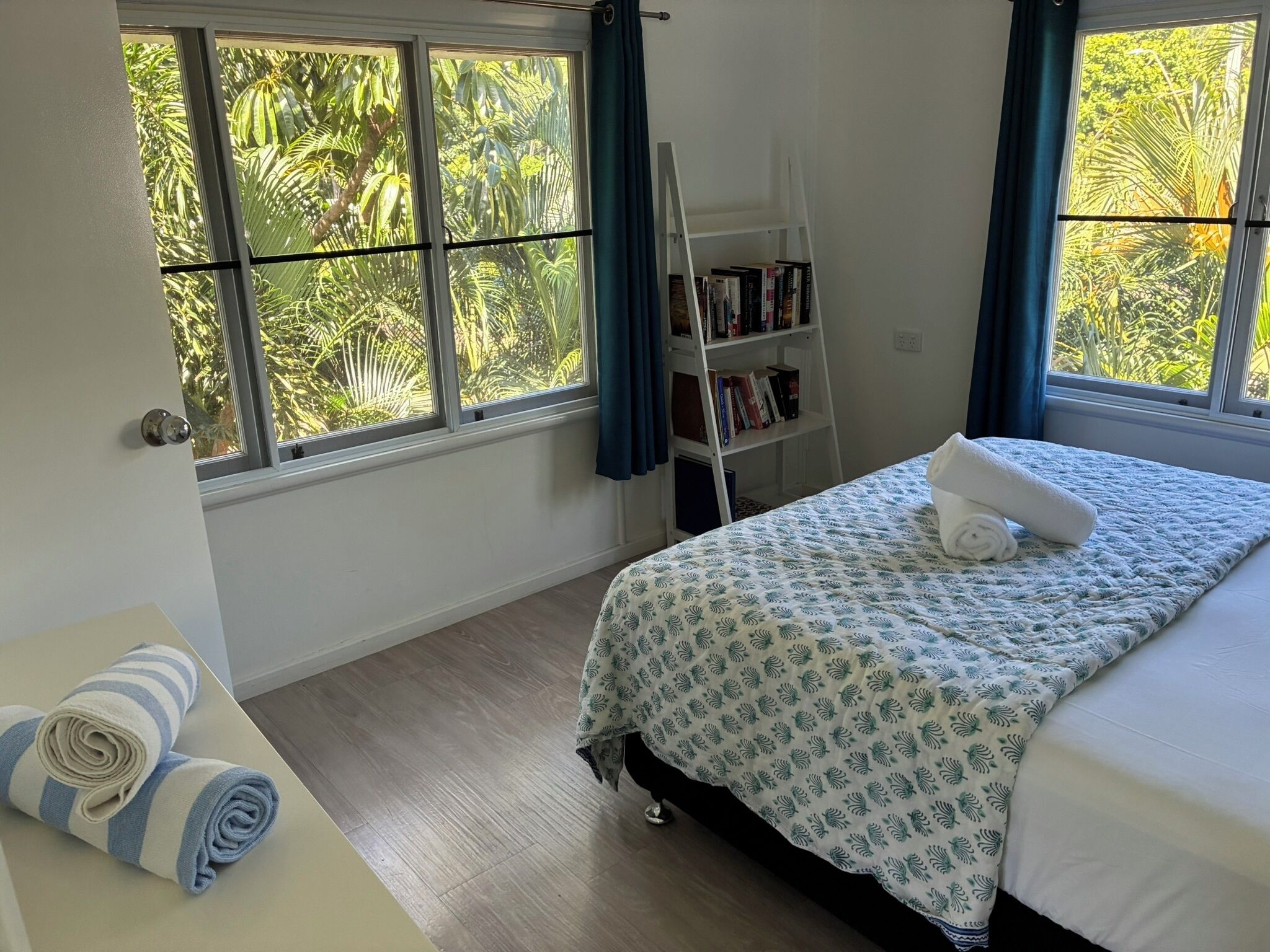 A bedroom with a bed, a bookshelf, and a window with a view of greenery.