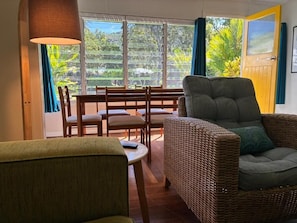 A room with a wicker sofa, a dining table with chairs, a lamp, and a view of greenery through large windows. - Kooyong Aprtments (Arcadia)