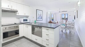 A modern kitchen with white cabinetry, a built-in microwave, and a dishwasher. There is a dining area with a table and chairs. - 2 Bedroom Unit with Beach & Golf at your doorstep! (Bargara)