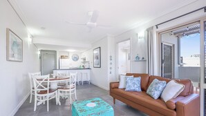 A modern living room with a brown leather sofa, white chairs, a kitchen area, and a ceiling fan. - 2 Bedroom Unit with Beach & Golf at your doorstep! (Bargara)