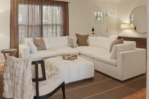 A modern living room with a sectional sofa, a small side table, a chair with a throw blanket, and a large window with curtains. - Real Home Near UT & Downtown - 3 Bed 2 Bath, Big Yard, Great Design (Austin)