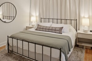 A neatly made bed with a metal frame, two bedside tables with lamps, a round mirror on the wall, and sheer curtains. - Real Home Near UT & Downtown - 3 Bed 2 Bath, Big Yard, Great Design (Austin)