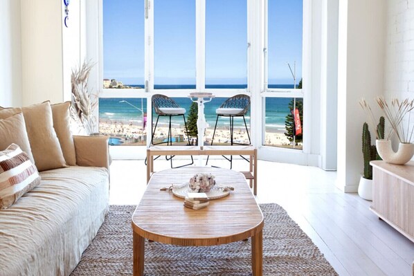 Ein Zimmer mit großem Fenster mit Blick auf einen Strand, einem Holztisch mit Dekoration, einer Couch und zwei Stühlen.