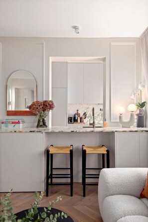 A modern kitchen with a white countertop, two wooden stools, a white sofa, and a large mirror. - Deluxe Bondi Suite - AC Laundry (Bondi Beach)