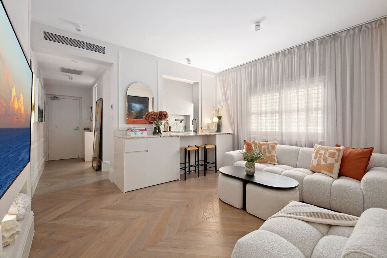 Ein modernes Wohnzimmer mit einer Couch, einem Couchtisch und einem Fernseher. Es gibt eine Küchenzeile mit einem Spülbecken und Hockern.