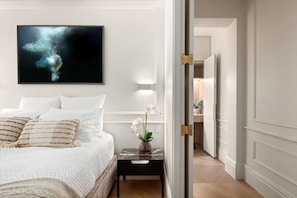 A bedroom with a bed, pillows, a nightstand with a vase, and a painting on the wall. - Deluxe Bondi Suite - AC Laundry (Bondi Beach)