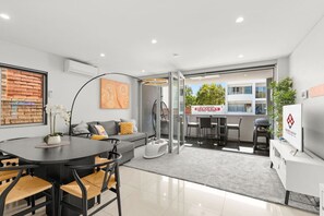 A modern living room with a sofa, dining table, and chairs, a kitchen area with a grill, and a large window with a view of the outside. - Stylish & Spacious Modern 1 BDR Balcony BBQ AC (Bondi Beach)
