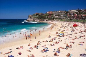 A crowded beach with people sunbathing and swimming, a red flag, and buildings in the background. - Stylish & Spacious Modern 1 BDR Balcony BBQ AC (Bondi Beach)