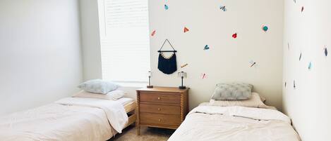 Une chambre avec deux lits simples, une commode en bois et une fenêtre avec des stores.