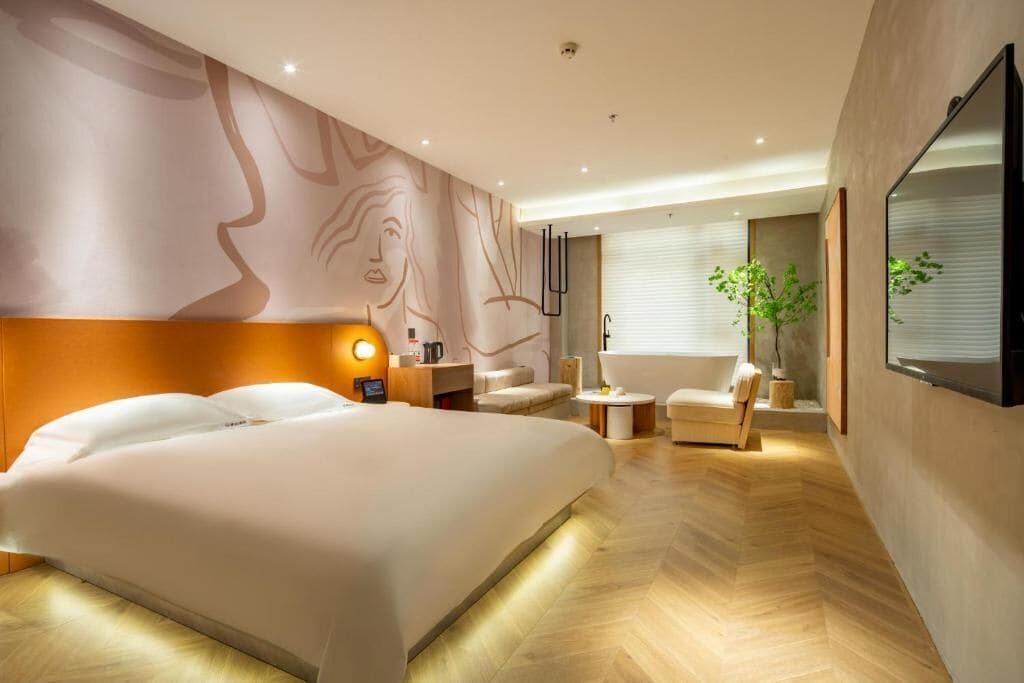 A modern hotel room with a large bed, a sitting area with a chair and a small table, a bathtub, and a large mirror on the wall.