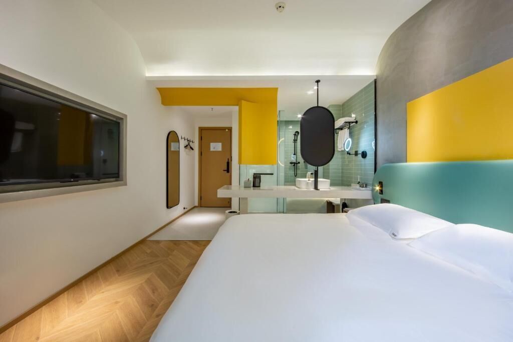 A modern hotel room with a large bed, a bathroom with a mirror and sink, and a window.