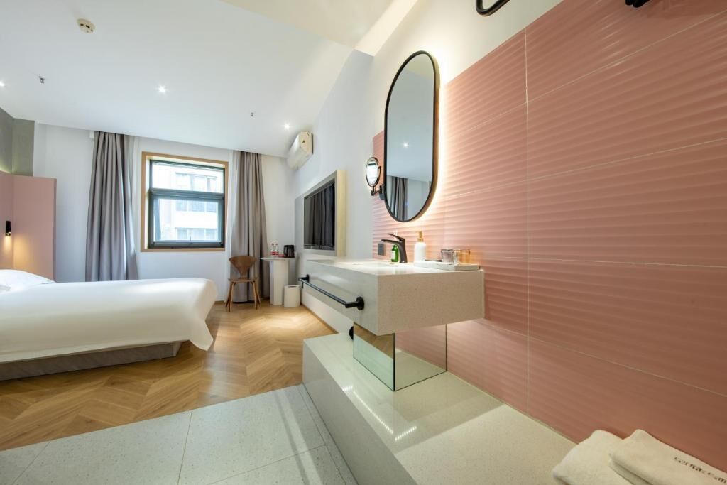 A modern hotel room with a bed, a vanity with a mirror, a television, and a window with curtains.