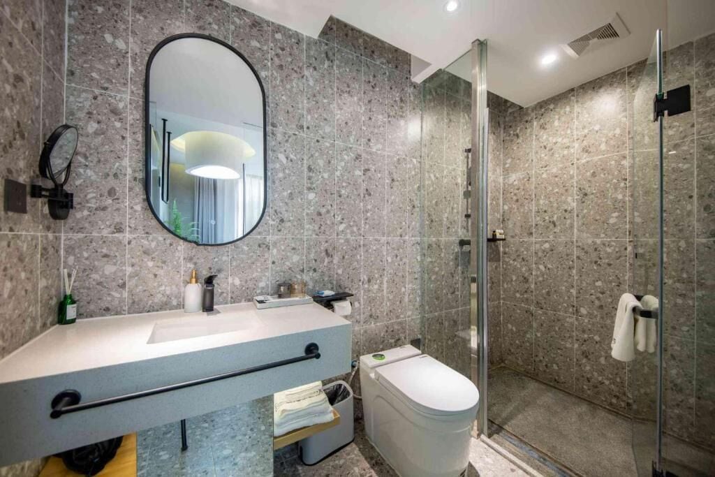 Een badkamer met een marmeren spatwand, een ronde spiegel, een toilet, een douchecabine en een handdoekhouder.