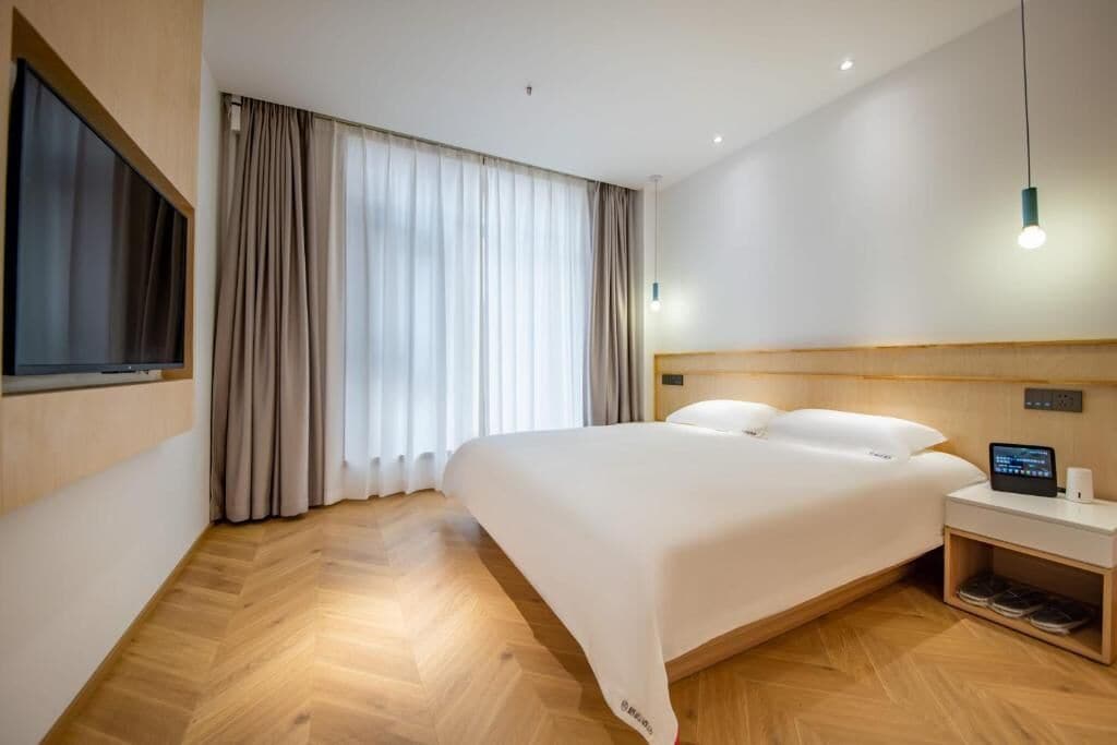 Una habitación de hotel moderna con una cama grande, un televisor montado en la pared, una mesita de noche con panel de control y suelo de madera.