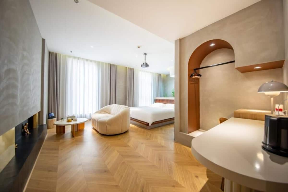 Una habitación de hotel moderna con suelo de madera en patrón de espiga, una mesa de centro redonda, un sofá, una cama y un escritorio con una lámpara.