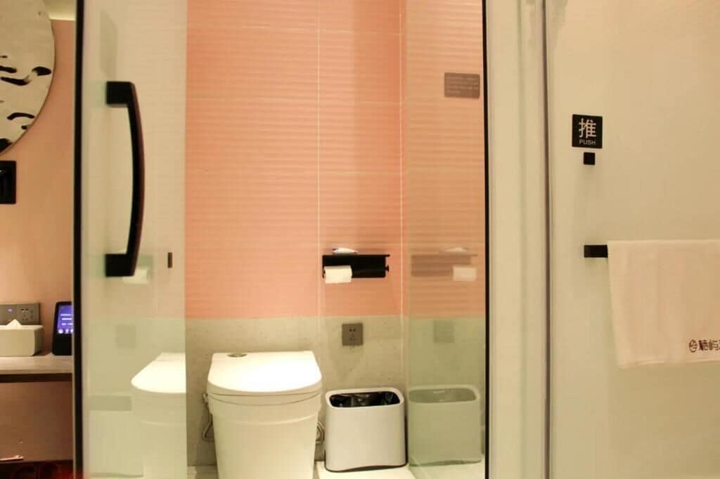 Un baño con inodoro, un cubo de basura, una toalla colgada de un gancho y un botón pulsador en la pared.